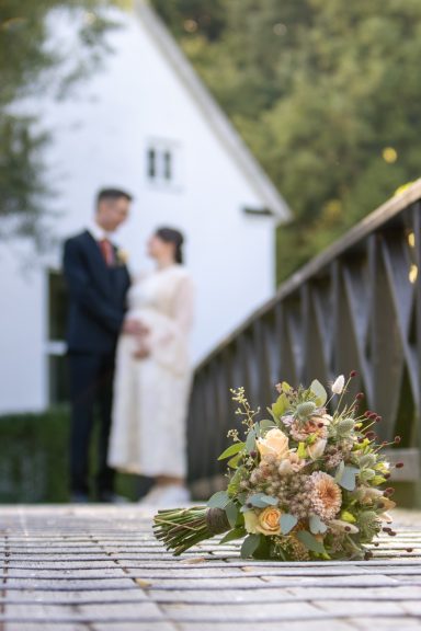 Detaljbild på brudbukett som ligger på en bro och brudparet syns ur fokus längre bort, bröllopsfoto av Anna Esmelöv