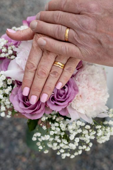 Detaljbild av brudens och brudgummens händer med vigselringar och med brudbuketten som bakgrund, bröllopsfotograf Anna Esmelöv