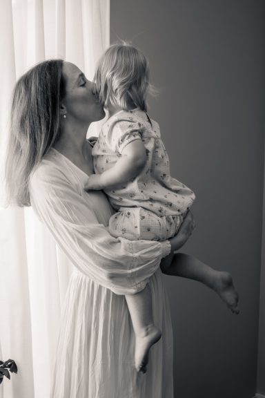 Svartvitt foto med avslappnad stil där mamma håller sitt barn inomhus vid ett fönster, familjefotograf Anna Esmelöv, Östergötland