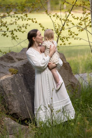 Mamma står lutad mot en sten i skogsbryn och håller barn i famnen, foto med fokus på mammor av Anna Esmelöv, Linköping