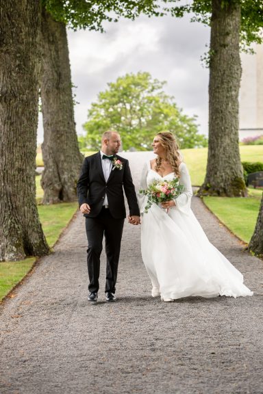Brudpar går i en allé av vackra träd och tittar kärleksfullt på varandra, Kuddby kyrka utanför Söderköping, foto av Anna Esmelöv