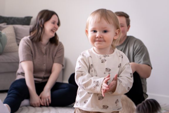 Barn leker med skrattande föräldrar i bakgrunden, familjefotografering inomhus med fokus på lifestyle och storytelling, Anna Esmelöv